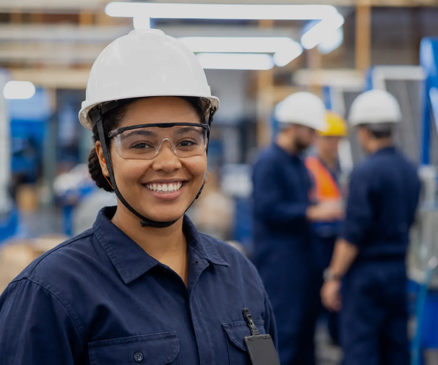 Production operator wearing a hard hat, safety glasses and earplugs, with her team working in the background